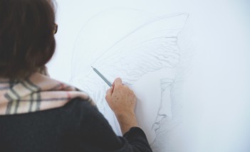Kobieta stojąca przy sztaludze i rysująca ołówkiem portret. 